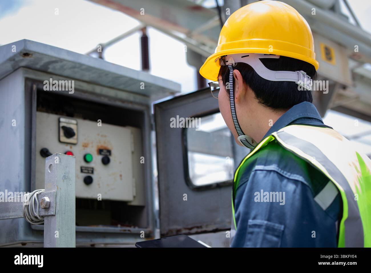 A power station engineer is working carefully inspecting electrical control panels wearing yellow safety helmet and reflective vest ensuring safety an Stock Photo