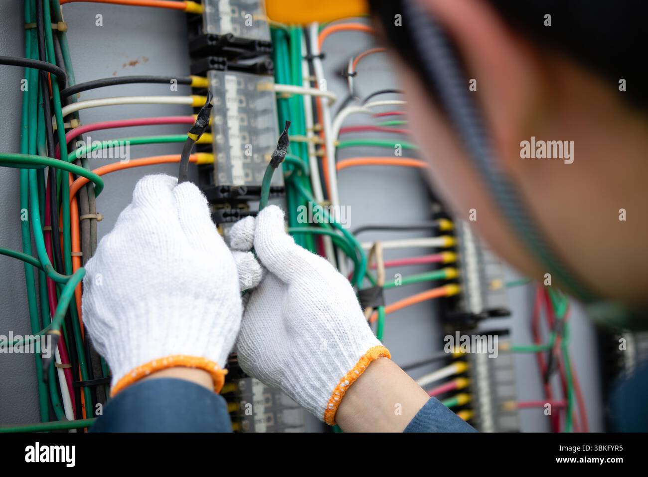 A power station engineer working with electrical wires wearing white gloves and safety helmet focused on repairing and maintaining system with colorfu Stock Photo