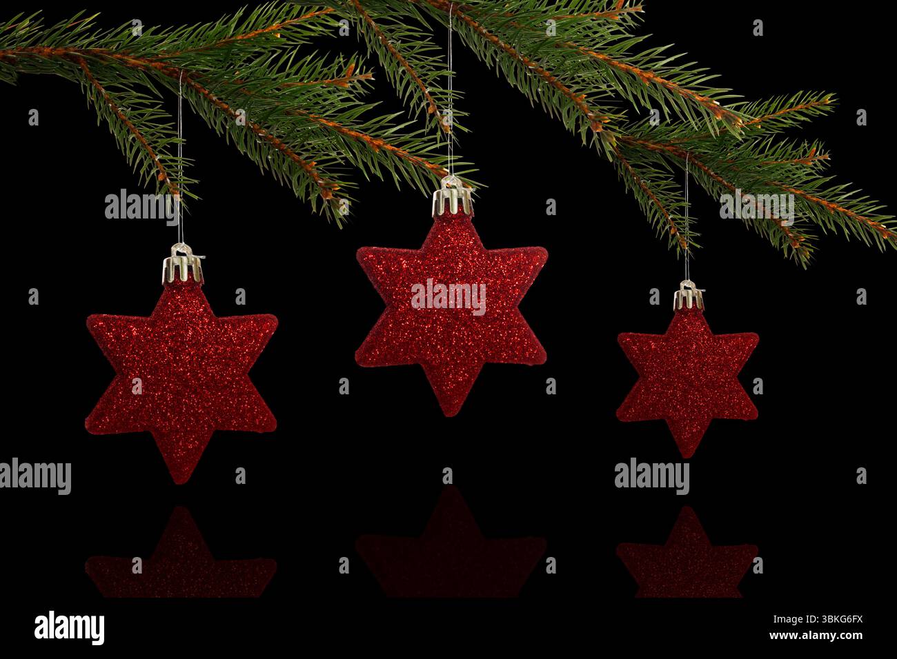 Three red glittery star ornaments are hanging on pine branch above black glossy surface, copy space Stock Photo