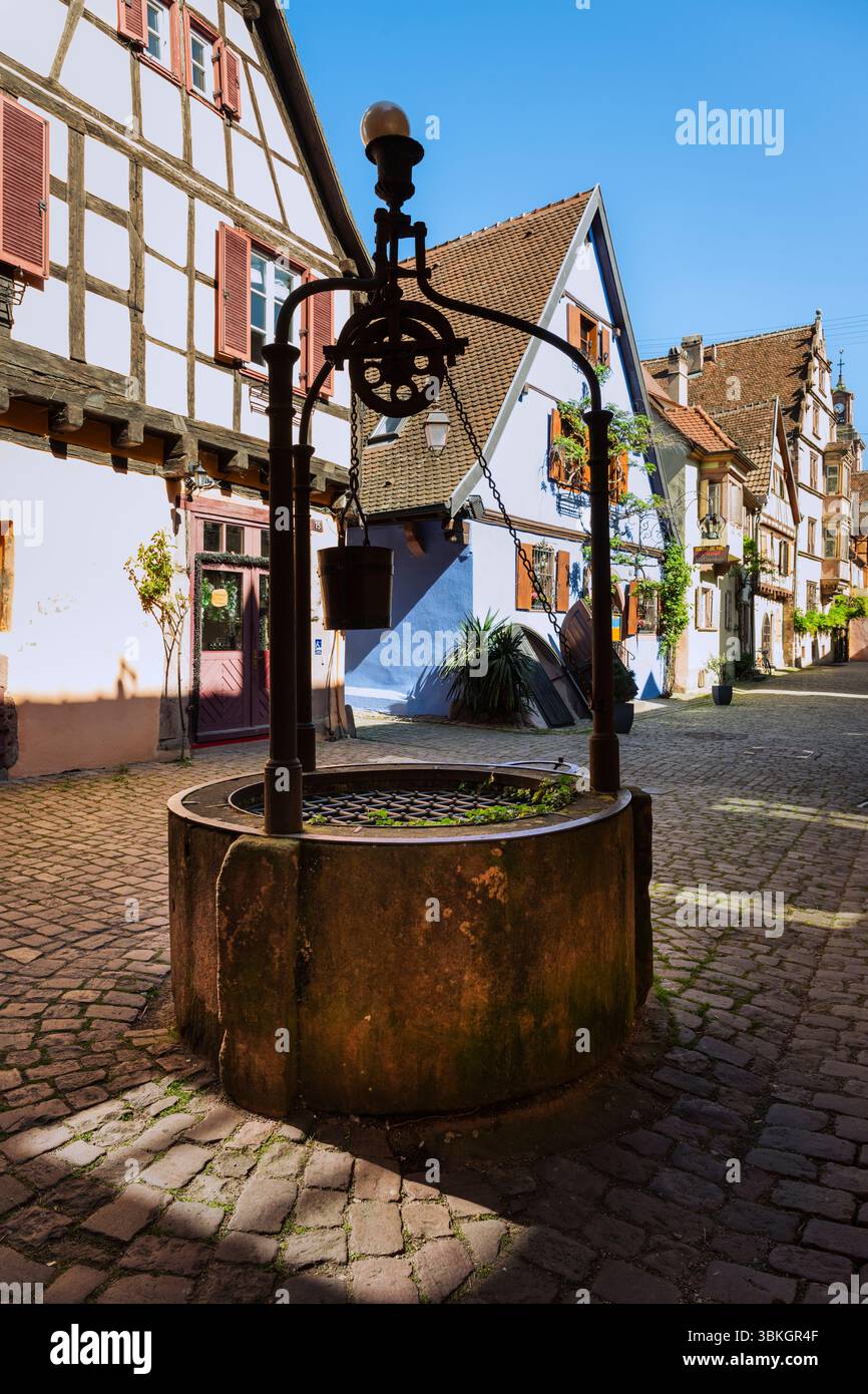Medieval well in front of half-timbered houses in the old town centre of Riquewihr in the morning sun, Alsace, France Stock Photo
