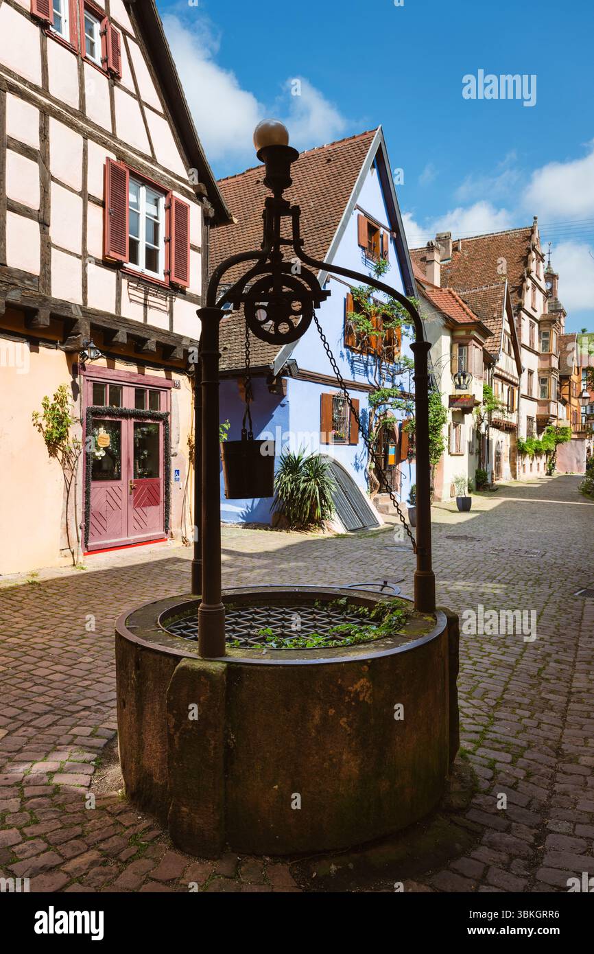 Medieval well in front of half-timbered houses in the old town centre of Riquewihr in the morning sun, Alsace, France Stock Photo