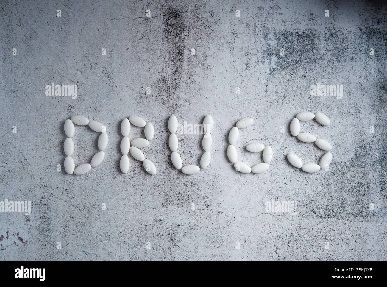 White oval pills arranged to form the word "drugs" on a textured, grey background. Concept of addiction, medicine, or illicit substances Stock Photo
