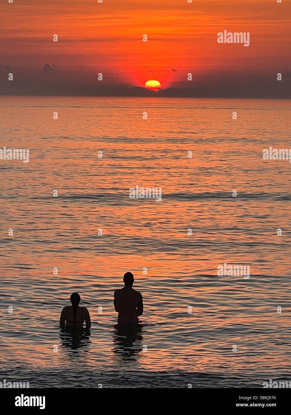 A couple enjoys a peaceful moment together in the calm waters as the sun rises, casting a warm glow over the ocean. - Smartphone Captured Stock Image