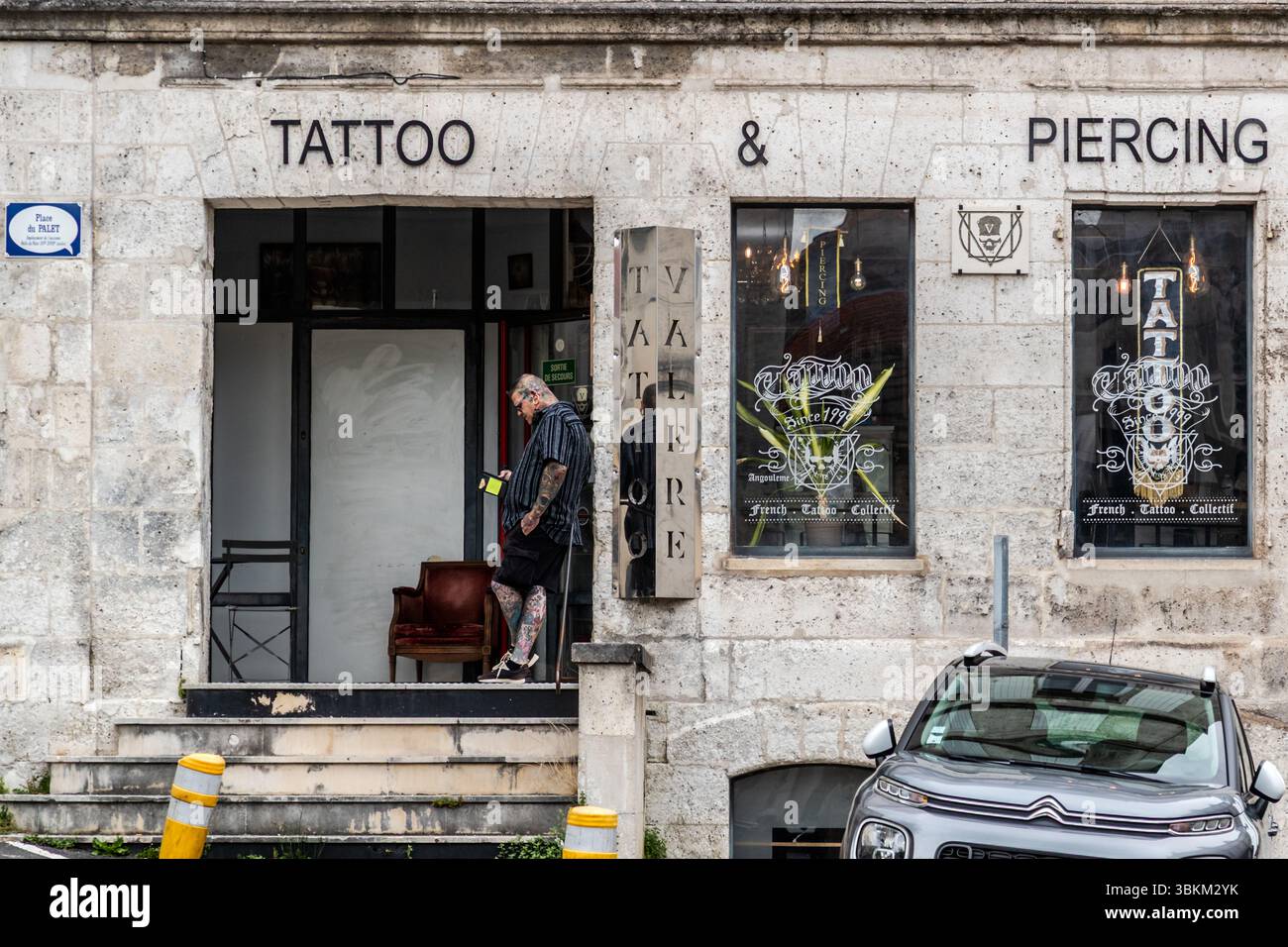 A heavily tattooed man stands in the doorway of a tattoo and piercing studio, Rue du Fort de Vaux, Angoulême, Nouvelle-Aquitaine, France. In the city, art and life merge: some people prefer to wear comics on their skin rather than hang them on their walls. The man, in shorts and a striped shirt, is busy with his mobile phone on the top step. Decorative windows and signs advertise the studio's services, the window lettering reads "French Tattoo Collectif". A street sign in the shape of a speech bubble names the square as Place du Palet. The city has been a UNESCO Creative City of Literature sin Stock Photo