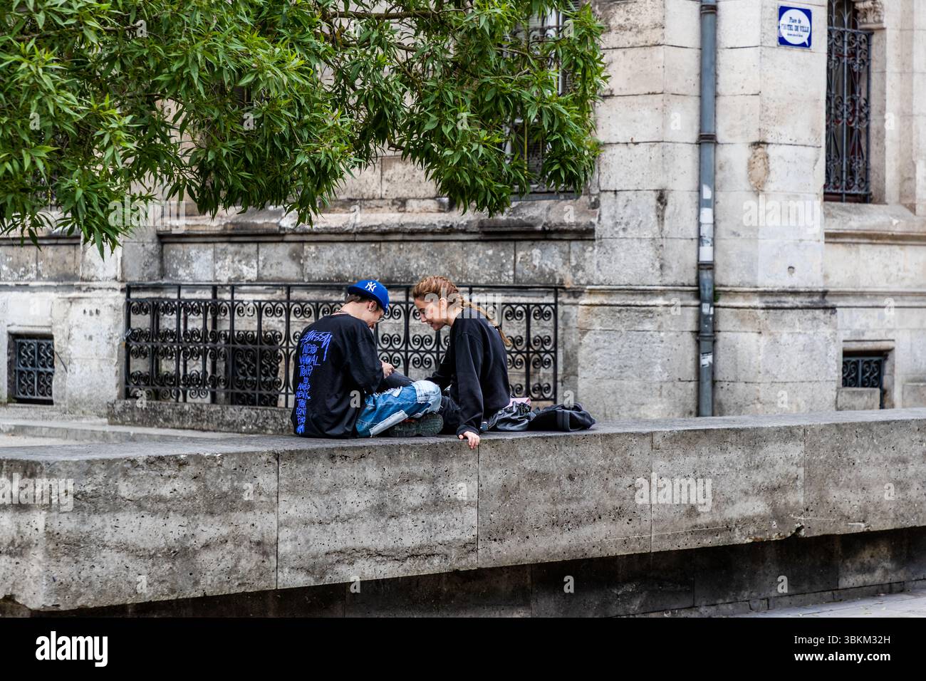 Everyday life in Angoulême: two young people sit on the wall outside the town hall and share a moment of digital togetherness, Rue de Genève, Nouvelle-Aquitaine, France. The street sign above them is shaped like a speech bubble and names the Place de l'Hôtel de Ville. Comics and the present merge. The young man wears a blue baseball cap and a black sweatshirt with a blue print, the young woman a black jumper and a braid. Both sit cross-legged on the stone wall, heads bent towards each other, in front of a wrought-iron grille. The city has been a UNESCO Creative City of Literature since 2019. Stock Photo