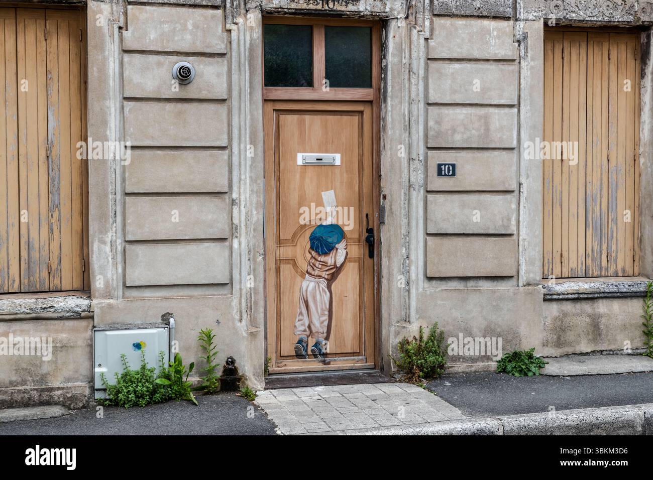 A painting on an outside door shows a little boy stretching in vain to put a letter in the real letterbox, Rue Léonard Jarraud, Angoulême, Nouvelle-Aquitaine, France. Even the letterbox becomes part of a tricky comic scene. The boy in a large blue cap, a striped jumper and baggy trousers stretches upwards and holds the letter high above his head. The painted wooden door belongs to house number 10 and stands between two shuttered doorways. The city has been a UNESCO Creative City of Literature since 2019. Stock Photo