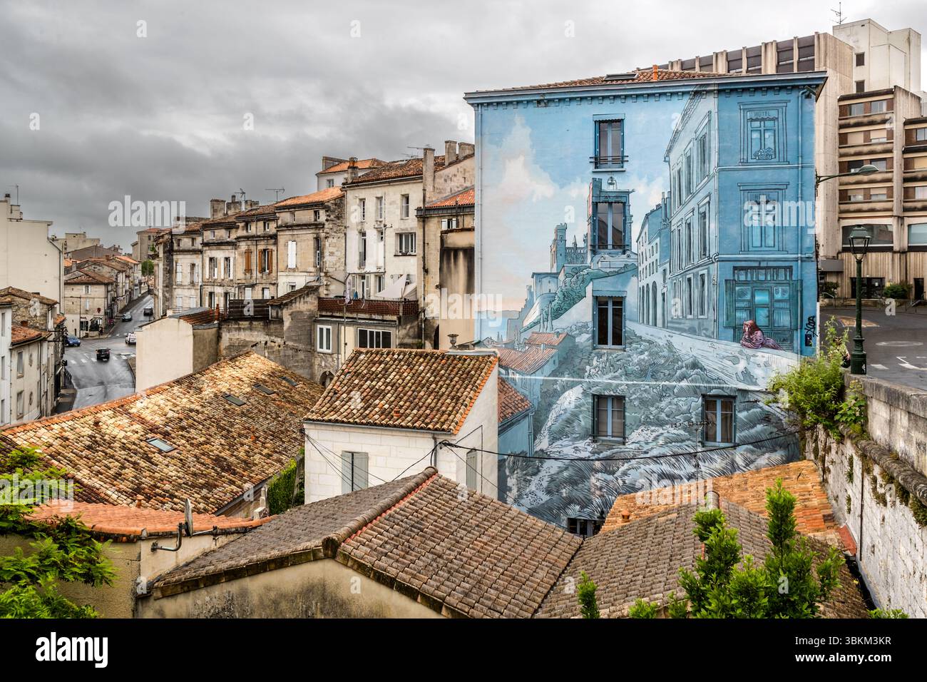 The mural "La Fille des remparts" (The Girl of the Ramparts) by Max Cabanes is an artistic deception that blends painting and the historic city walls, Rue de la Corderie, Angoulême, Nouvelle-Aquitaine, France. In it, a woman leans on a painted city wall that continues into the original city wall. The 120-square-metre trompe-l'œil dates from 2004. Its blue tones recall Béziers in the south of France, the comic artist's home town. Cabanes won the Grand Prix of the city's International Comics Festival in 1990. The blue gable rises above red-tiled rooftops, and a street lined with old townhouses r Stock Photo