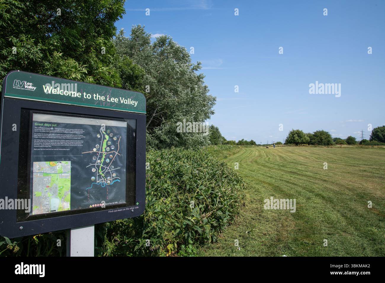 Welcome sign at Lee Valley Park with a mown grass trail, summer trees, and clear blue sky in the background. Stock Photo