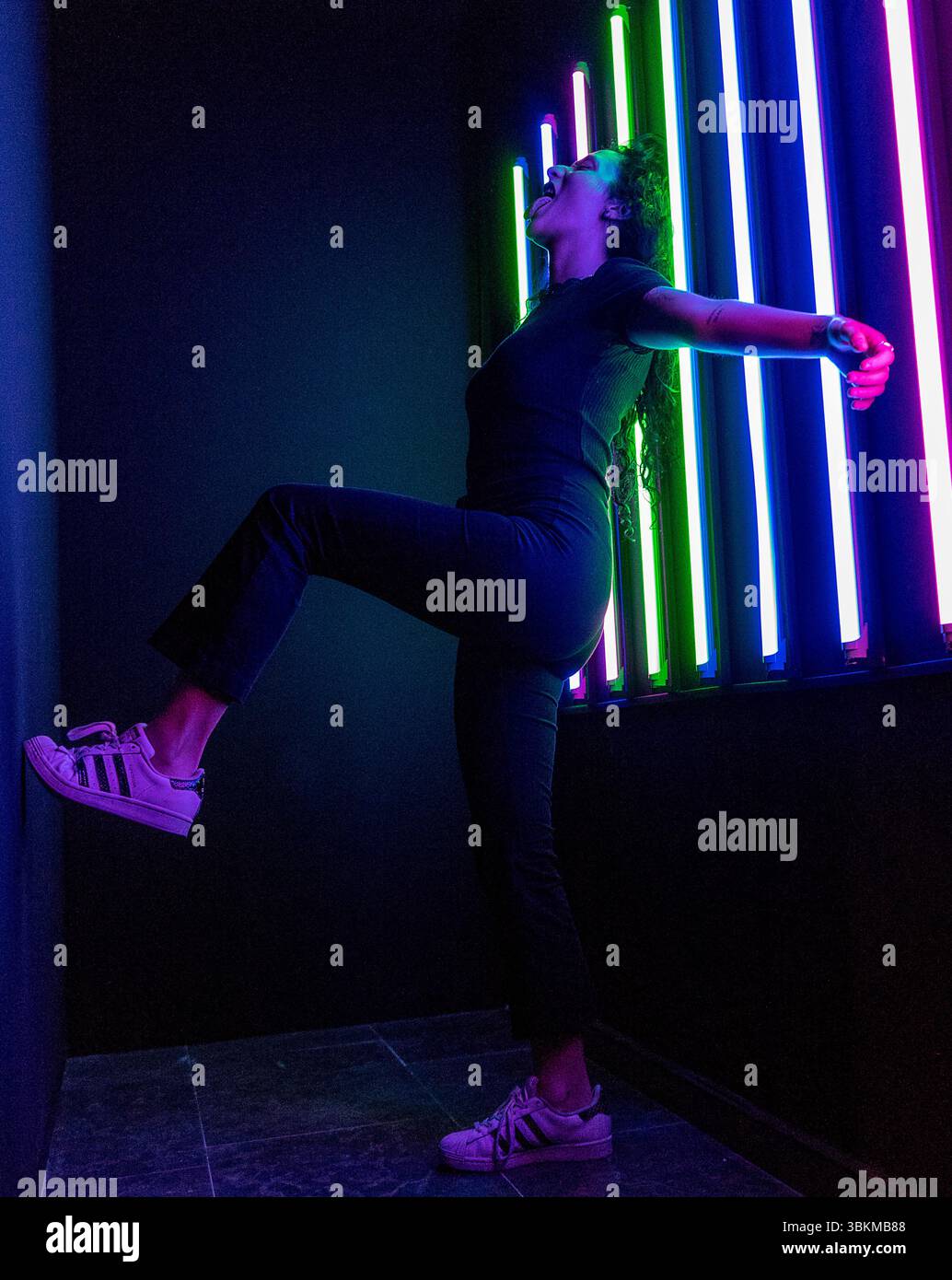 A woman in a dynamic pose standing in a dark room lit by vibrant neon lights, creating a striking contrast of colors and shadows. The image captures a Stock Photo