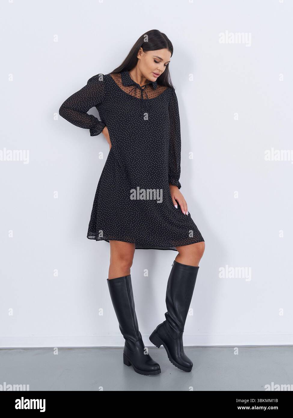 Dark-haired woman in knee-high black boots and sheer-sleeved dress poses against white wall, looking down. The dress has a delicate, intricate pattern Stock Photo