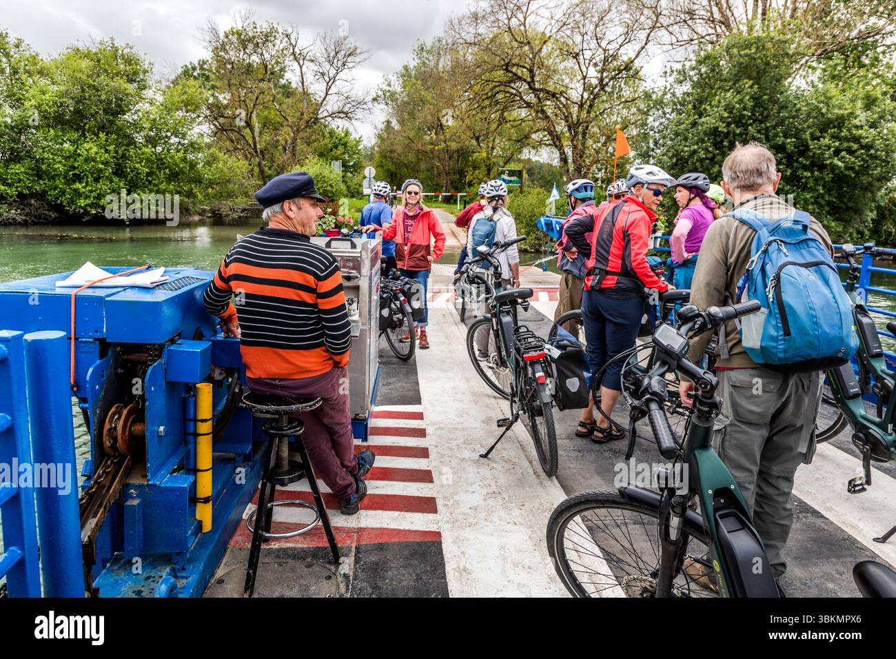 A group of cyclists crosses the Charente by ferry. They stand with their bicycles on a ferry in Chaniers, France. The ferryman, resting on his chair, steers the ferry. The scene shows a relaxed atmosphere and the joy of cycling in nature. Nouvelle-Aquitaine, France Stock Photo