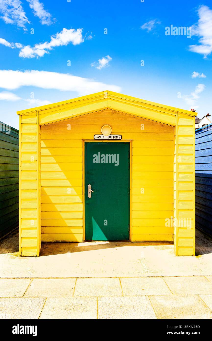 Bright yellow beach hut with a green door under a vibrant blue sky with white clouds, surrounded by other colorful huts Stock Photo