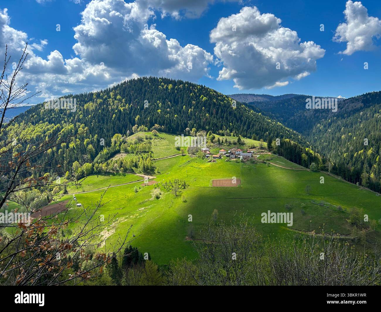 A scenic mountain village nestled in vibrant green hills and dense forest, under a dramatic sky with fluffy clouds casting shadows across the landscap - Smartphone Captured Stock Image