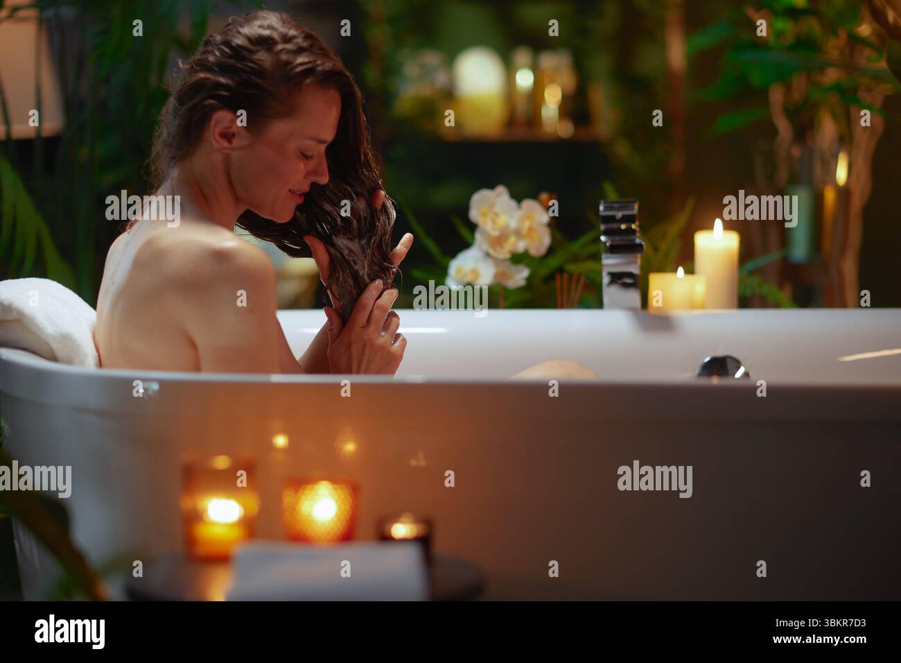 A woman with wet, dark hair in profile, washing/conditioning in a dimly lit bathtub. Surrounded by green plants, orchids, & candles, it creates a sere Stock Photo