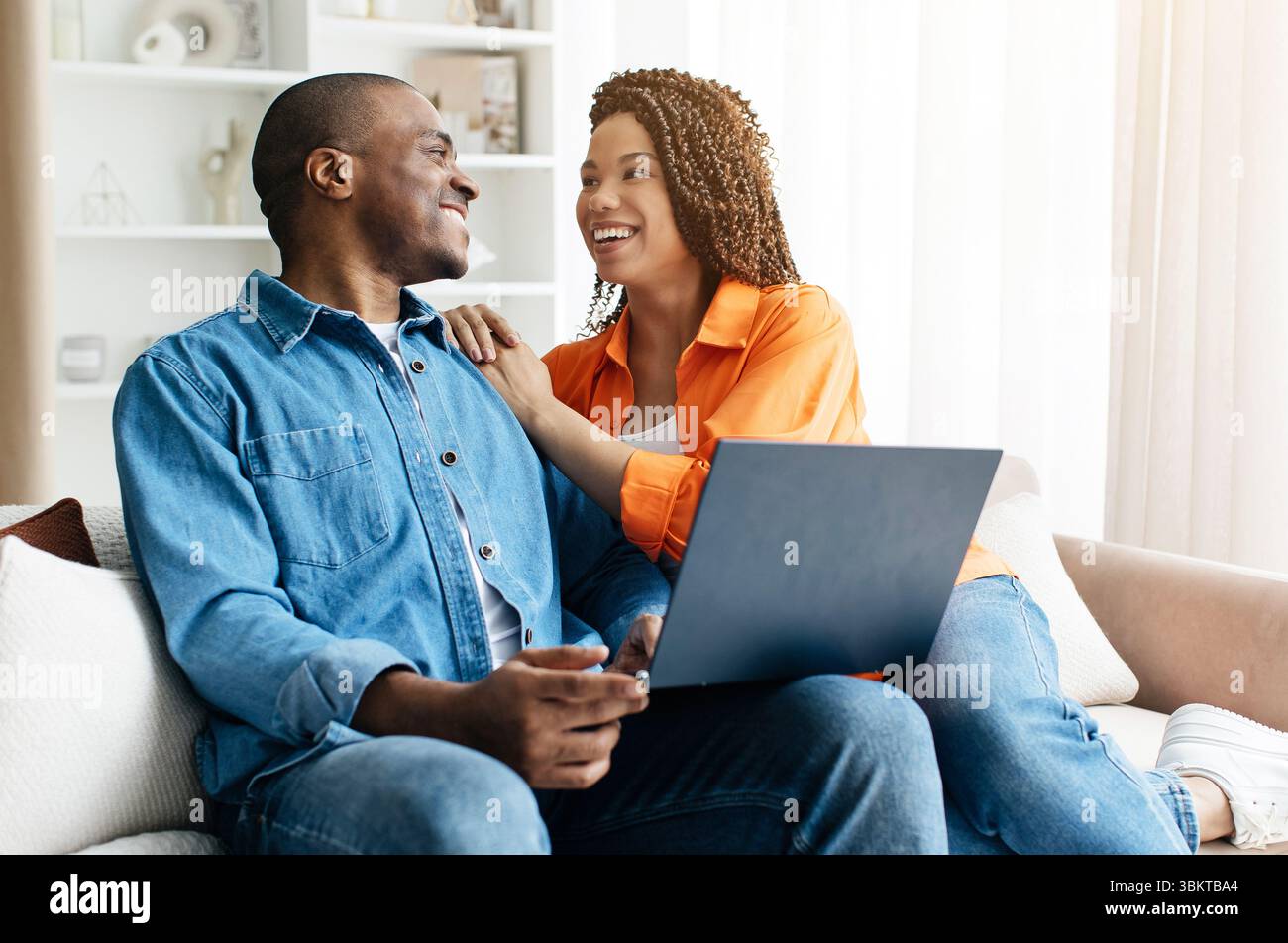 Black couple smiling at each other while sitting on couch with laptop Stock Photo