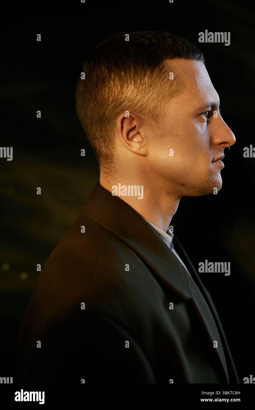 Vertical shot of Caucasian young adult man standing in profile view against dark background, showing short hair and clean facial features in studio Stock Photo