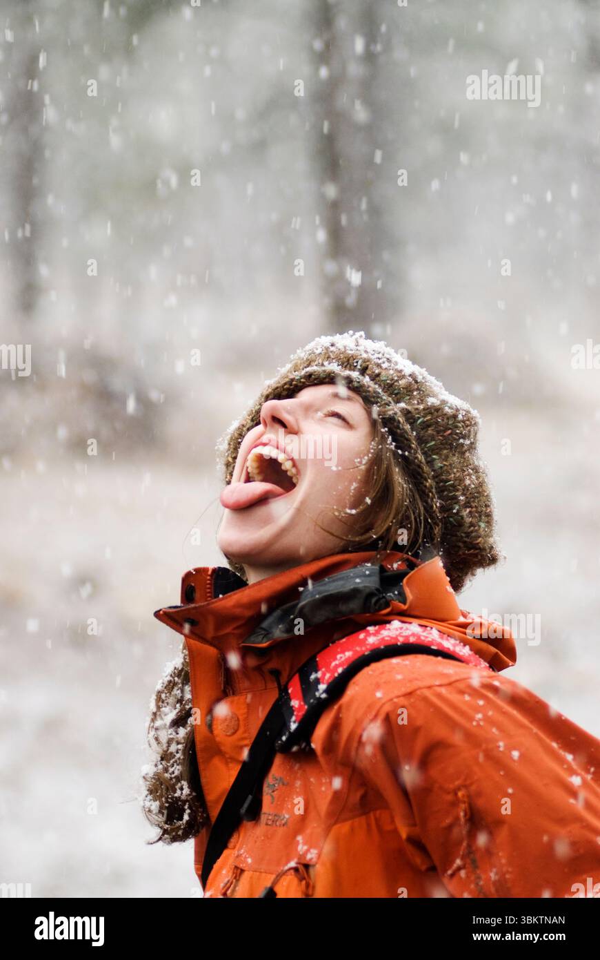 A young woman in an orange jacket laughs and smiles as she tries to catch snowflakes on her tongue during a winter storm in Flag Stock Photo