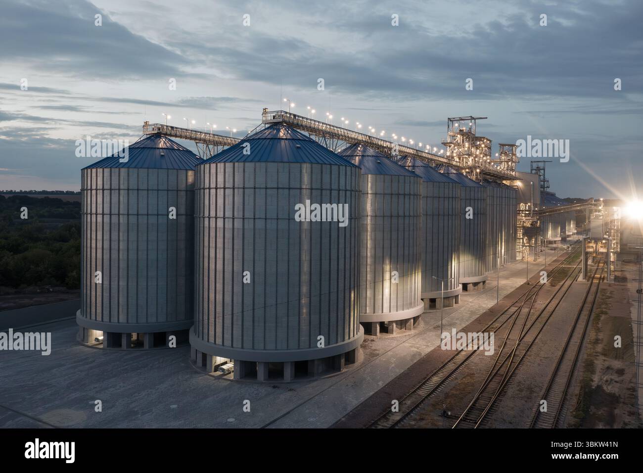 Night view of modern grain silos and elevator facility with railway, illuminated for 24 hour agriculture, export, and logistics operations. Stock Photo