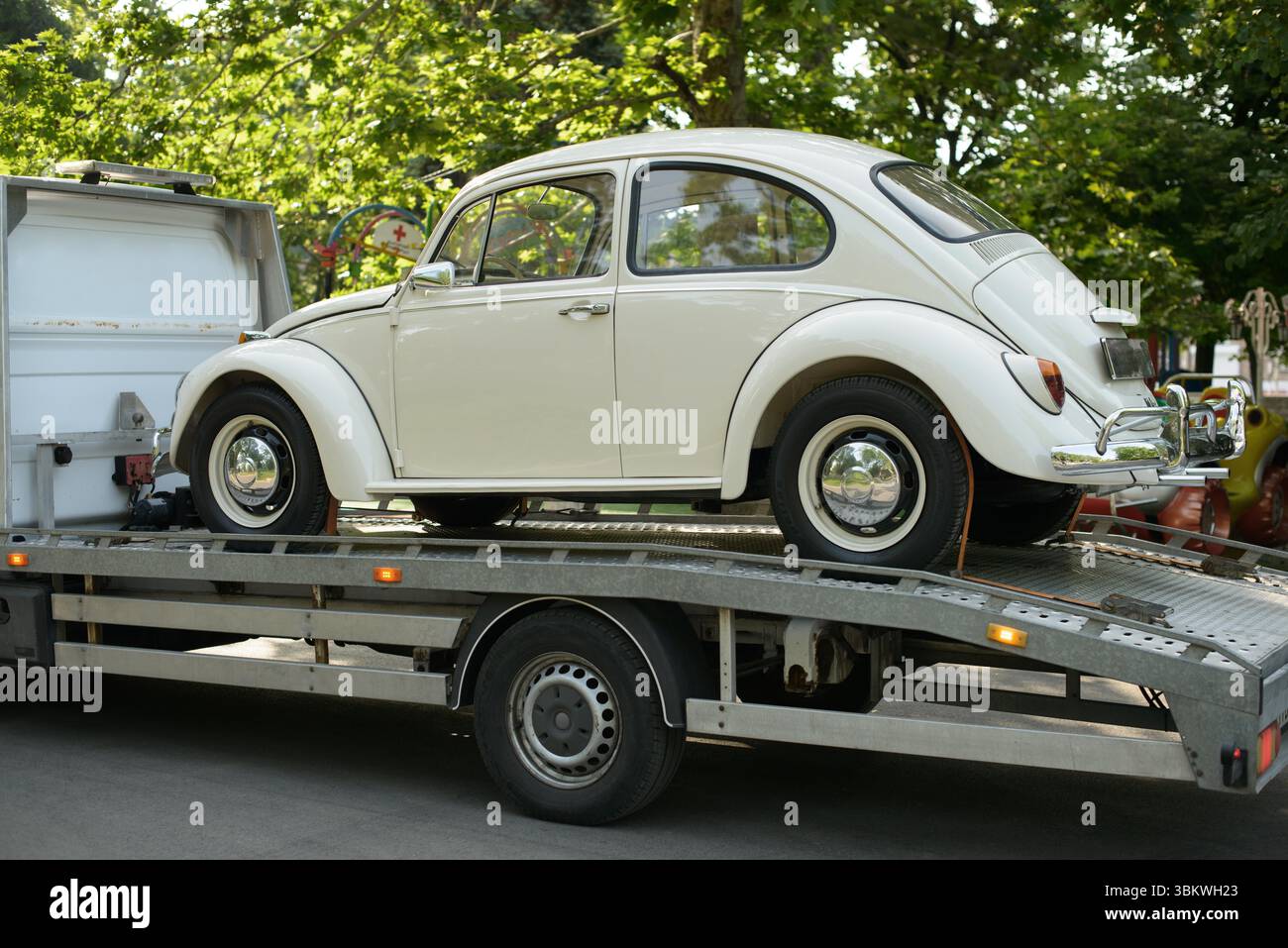 A cream-colored vintage two-door coupe sits on a flatbed transport truck, chrome hubcaps and slim whitewall tires glinting beneath leafy summer trees Stock Photo