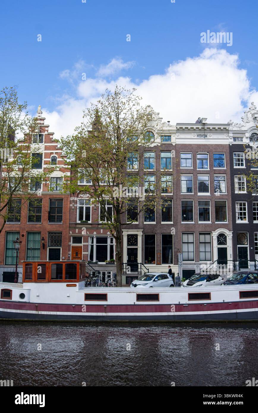 Daily life on an Amsterdam canal with historic houses, a houseboat, and a person walking. Stock Photo