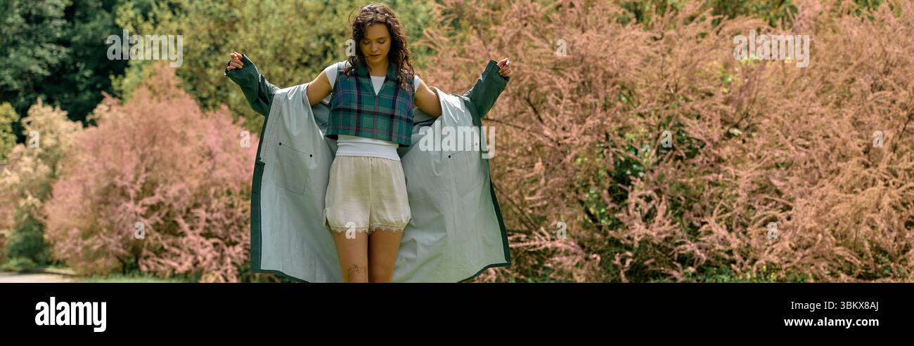 Curly-haired woman showcases a trendy raincoat while enjoying the outdoors in summer, banner Stock Photo
