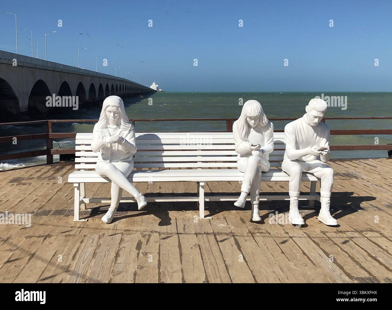 White sculpture of tourists using cell phones on a bench in Progreso, Mexico—playful seaside art capturing modern vacation life. Stock Photo