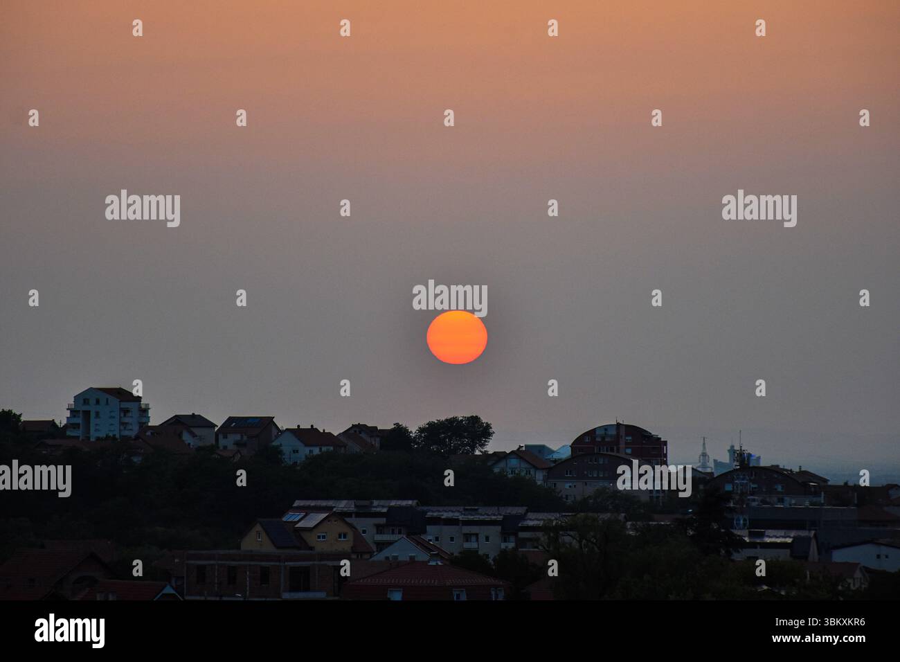 Glowing golden sun sets behind cityscape, with clear sky fading from deep orange through blue to soft bluish gray Stock Photo