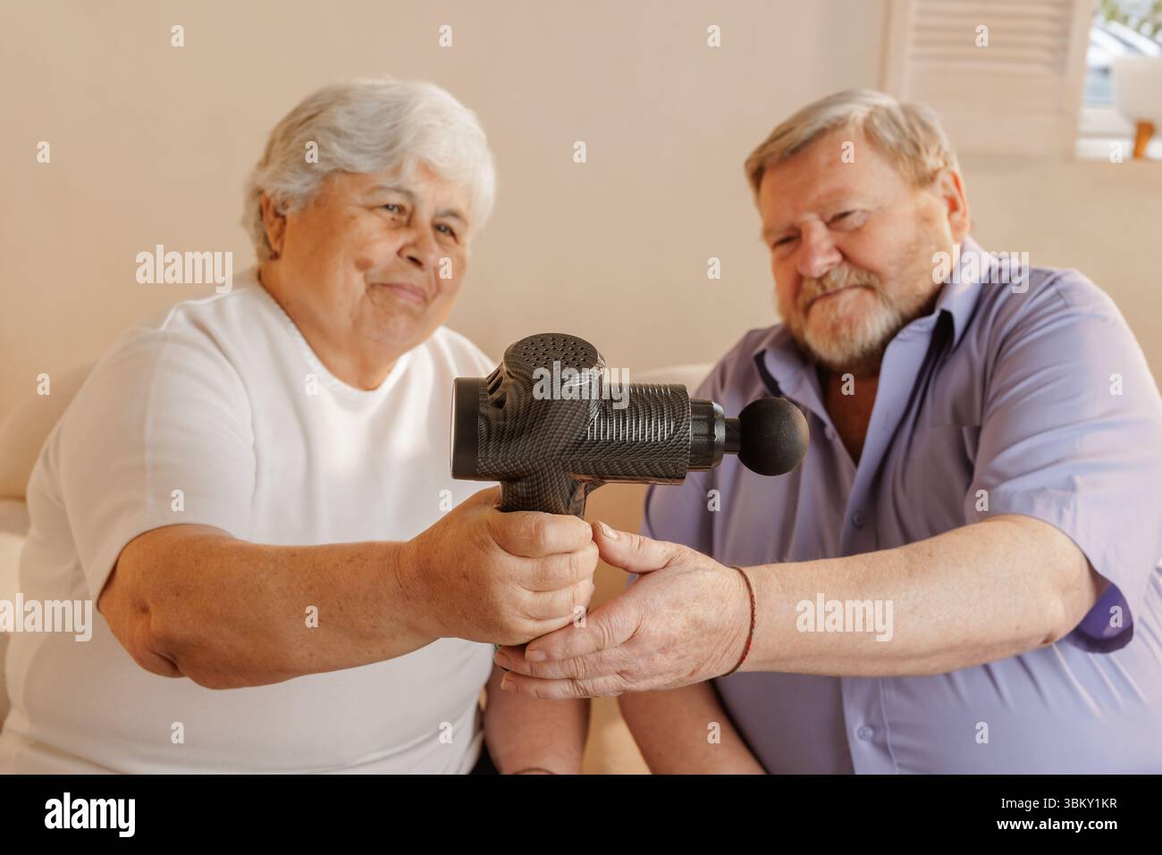 Elderly couple using a handheld electric massager at home for muscle pain relief wellness and comfortable self care routine Stock Photo