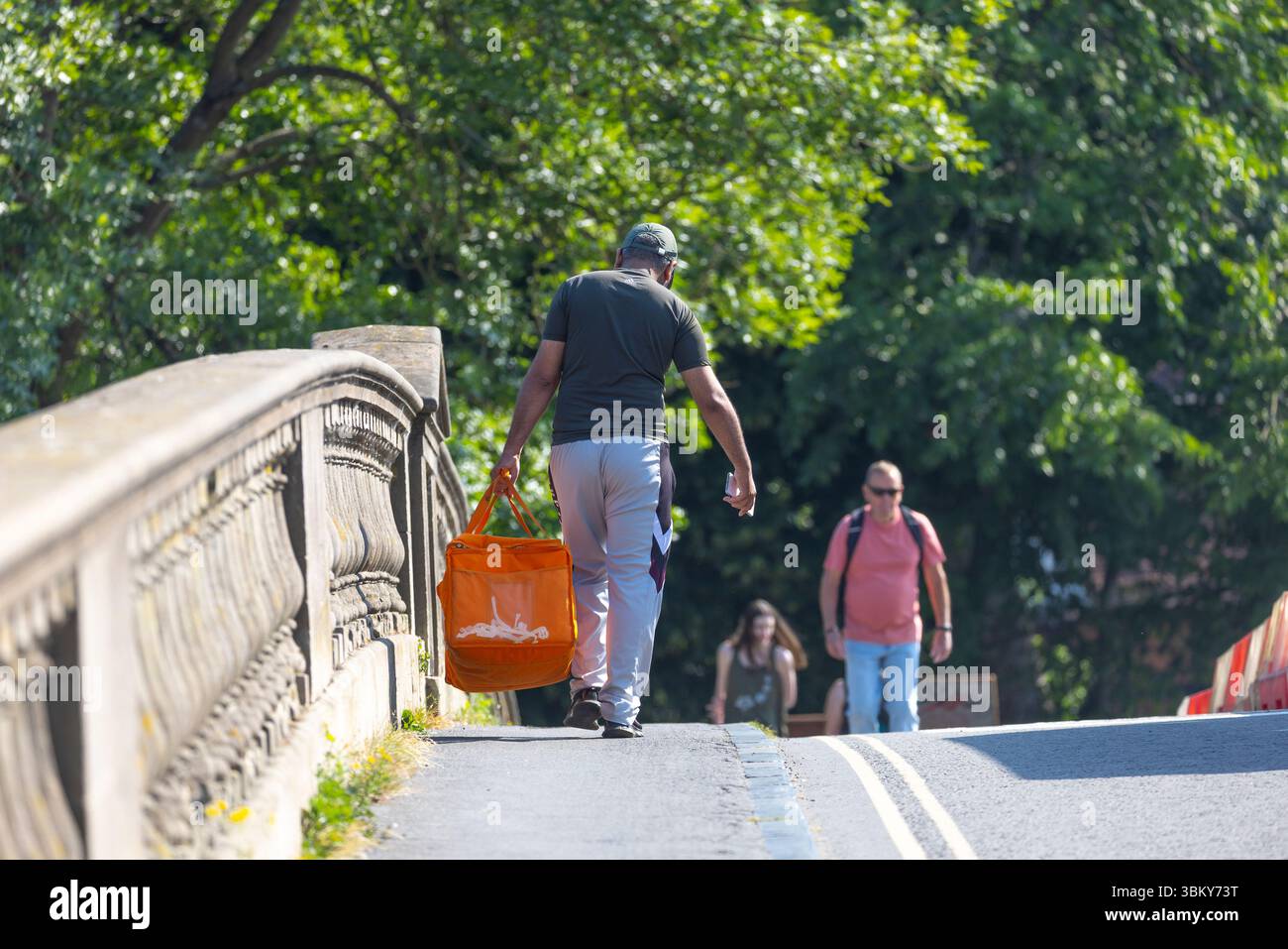 Food delivery man carrying an orange insulated bag on foot walking over a bridge in a UK town in the summer. Stock Photo
