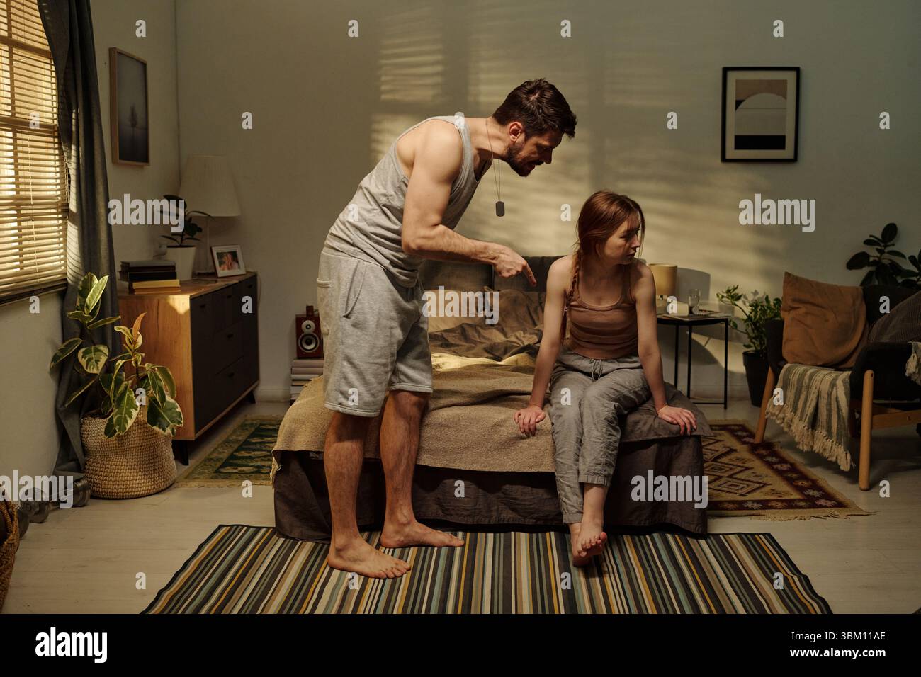 Man standing and gesturing angrily toward woman sitting on bed looking away, illustrating family conflict in domestic bedroom setting Stock Photo