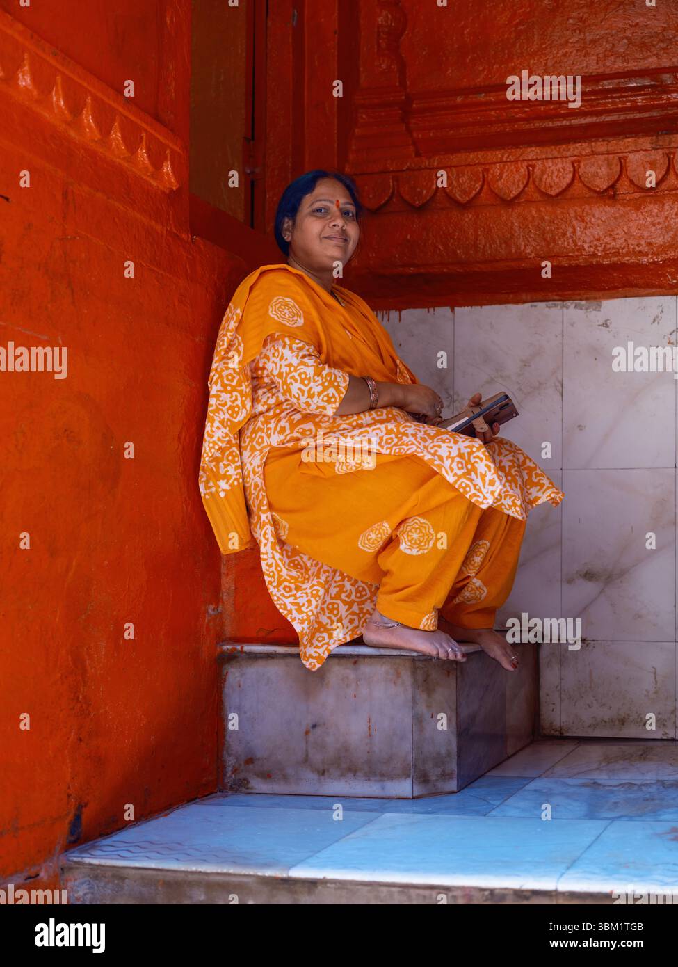 India, Varanasi, 16 Mar 2025. Woman in saffron salwar kameez sits barefoot in a red-painted temple alcove, holding a devotional booklet during a midda Stock Photo