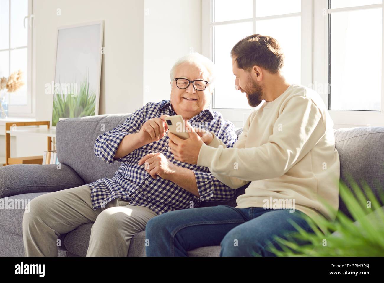 Senior father sitting on sofa at home with his adult son and using smartphone. Stock Photo