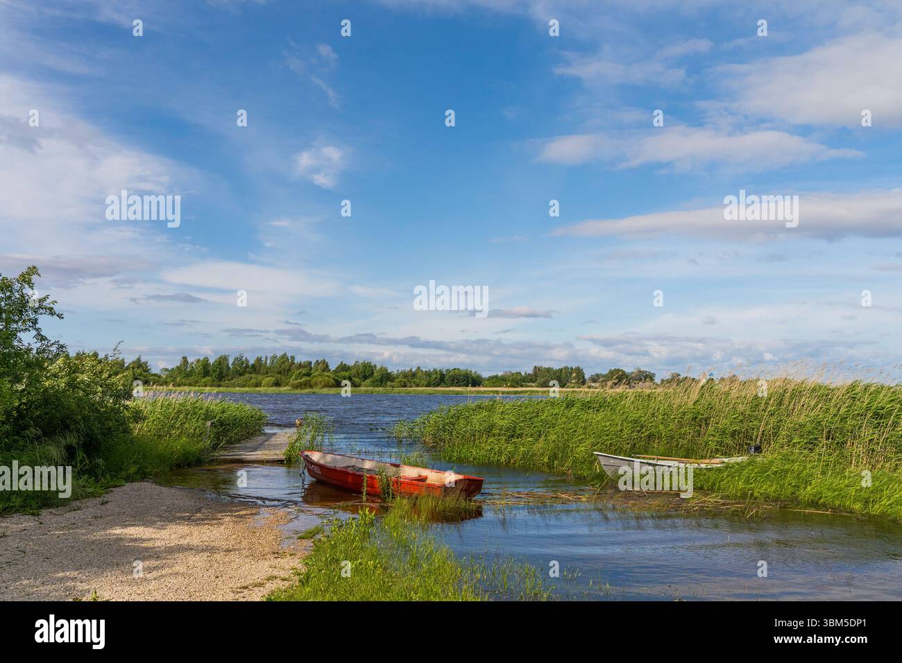 Red boat on the Estonian shore of Lake Peipus in Podmotsa, with young reeds, blue sky, and a distant view of Russia across the water. Stock Photo