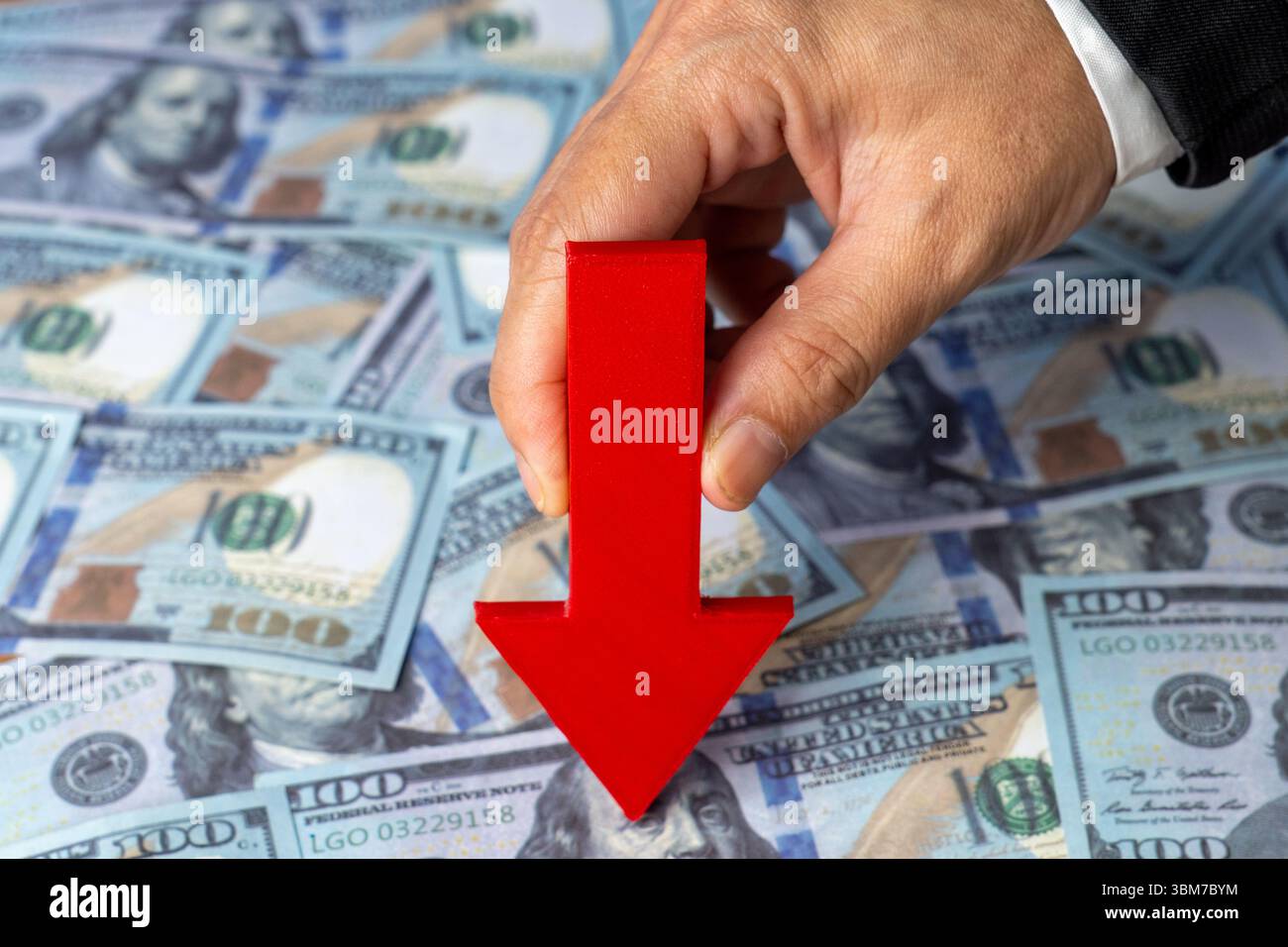 A hand holds a red arrow pointing downwards against a background of scattered US one-hundred dollar bills. Financial downturn, economic loss Stock Photo