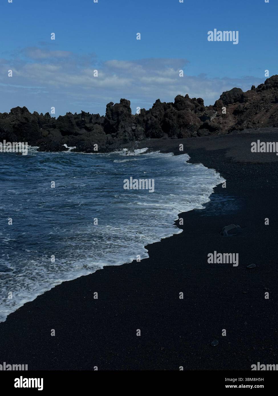 Black sand beach with gentle waves, rocky cliffs in the background, and clear blue sky. Serene coastal scene featuring a unique volcanic black sand. - Smartphone Captured Stock Image