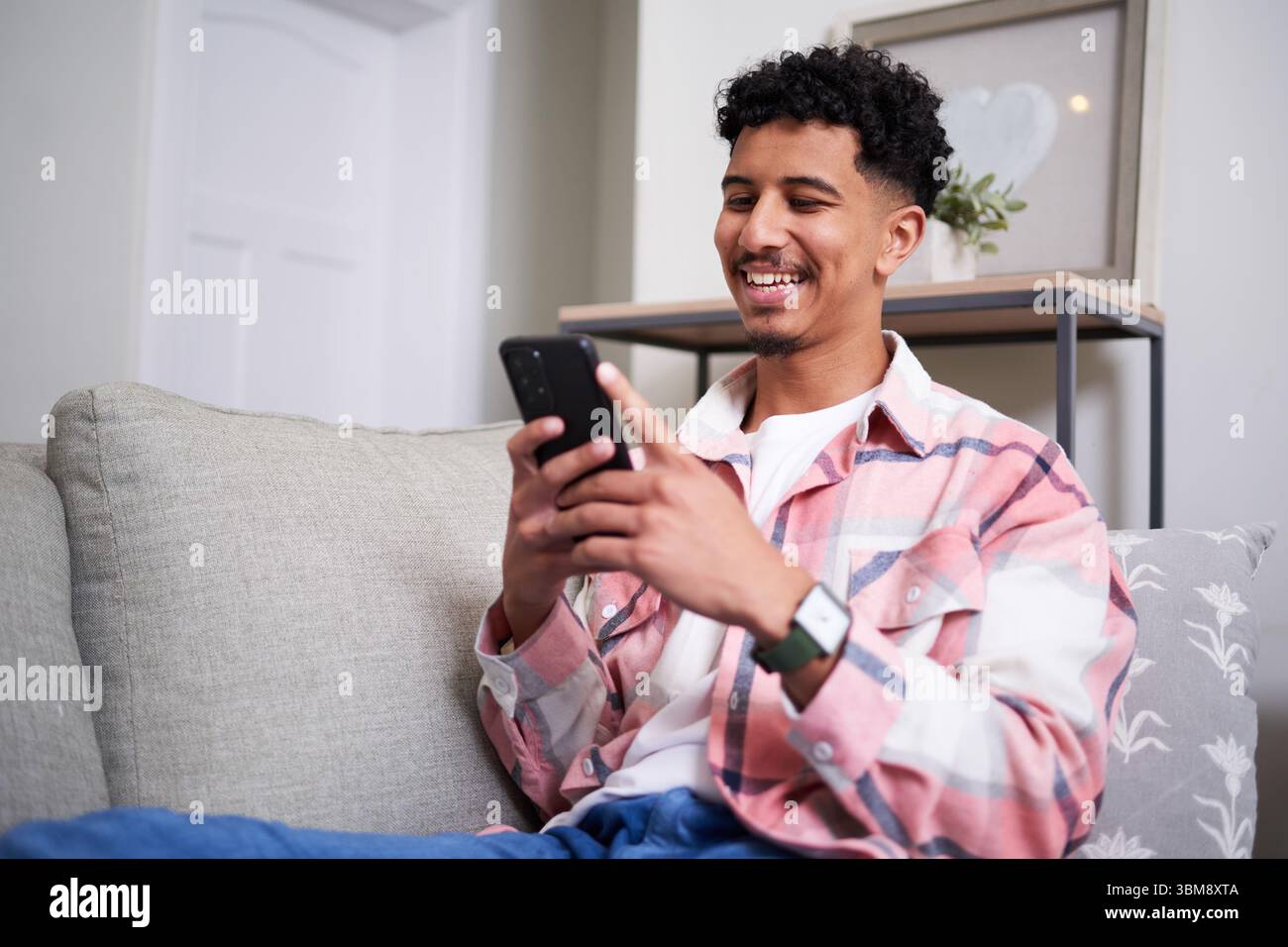 Smiling Man Relaxing at Home While Using Smartphone on Couch Stock Photo