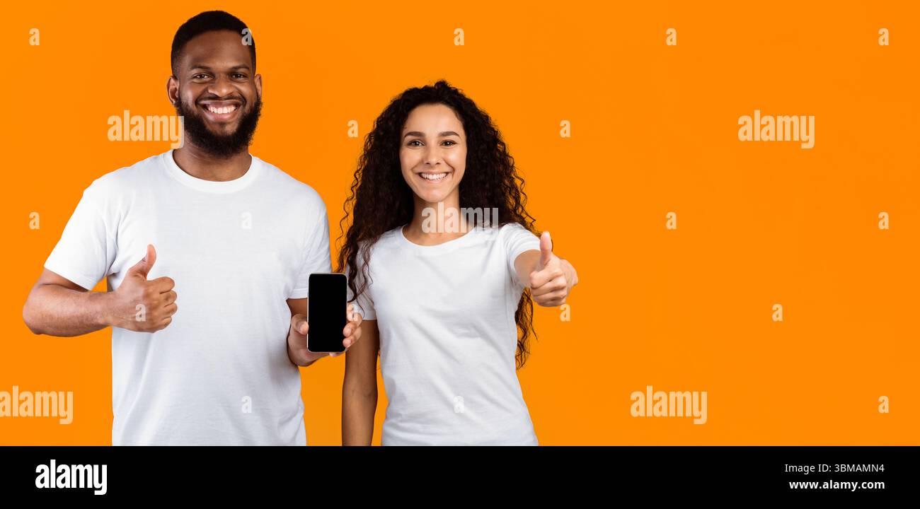 Multiethnic Couple Showing Cellphone Screen Gesturing Thumbs-Up Over Yellow Background Stock Photo