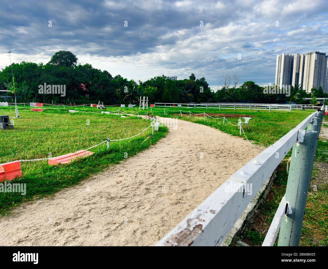 Wide shot of an empty horse riding field under an open sky, with a building in the background. A wooden fence appears in the left foreground - Smartphone Captured Stock Image