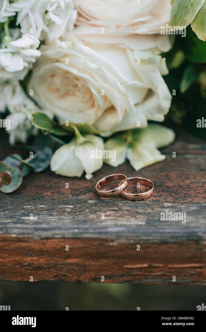 Roses gold wedding bands resting on rustic wooden bench beside ivory rose bouquet Stock Photo