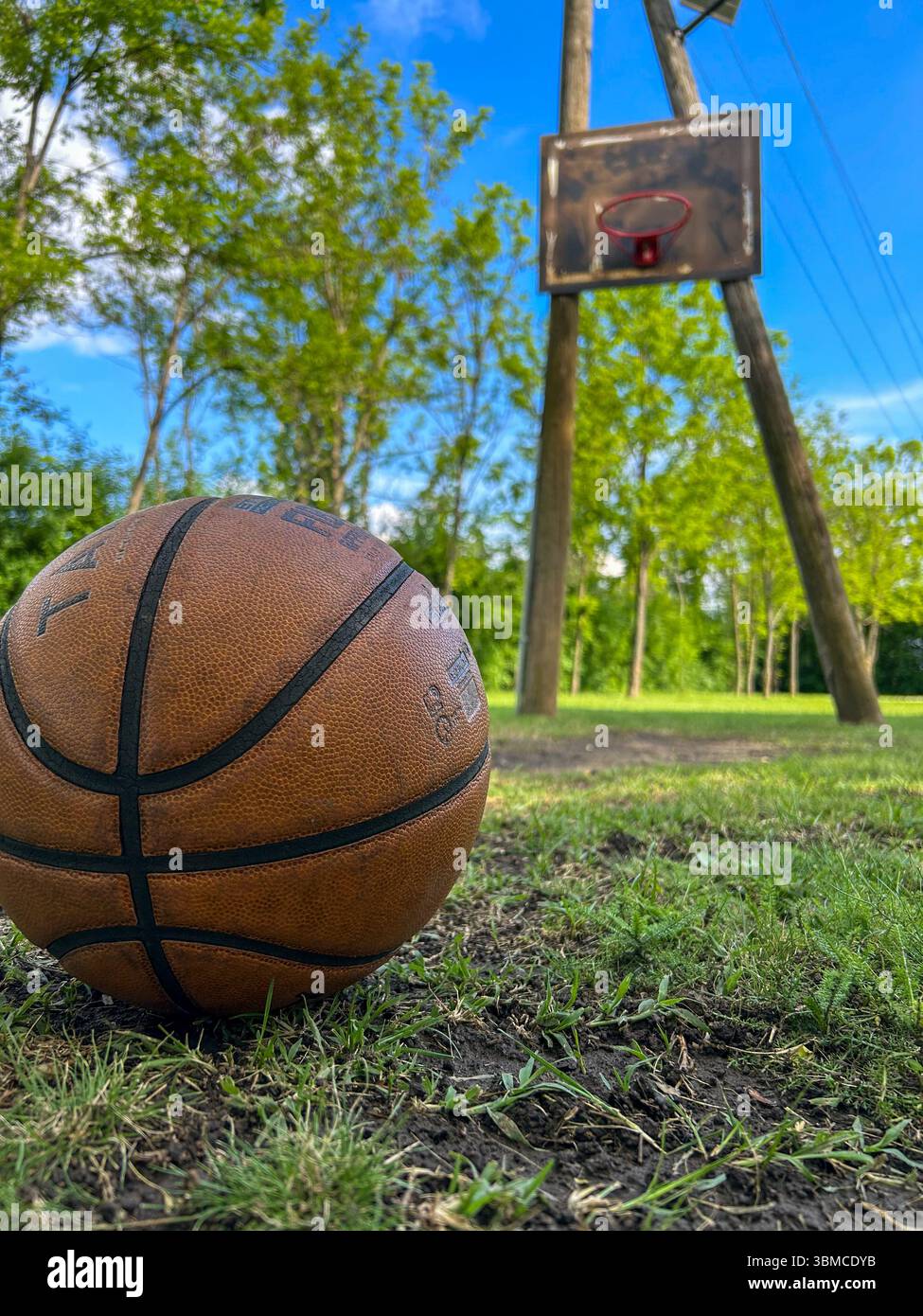 Close-up of a worn basketball on grass with an old wooden outdoor hoop in the background, set against a bright blue sky and green trees. Rural sports - Smartphone Captured Stock Image