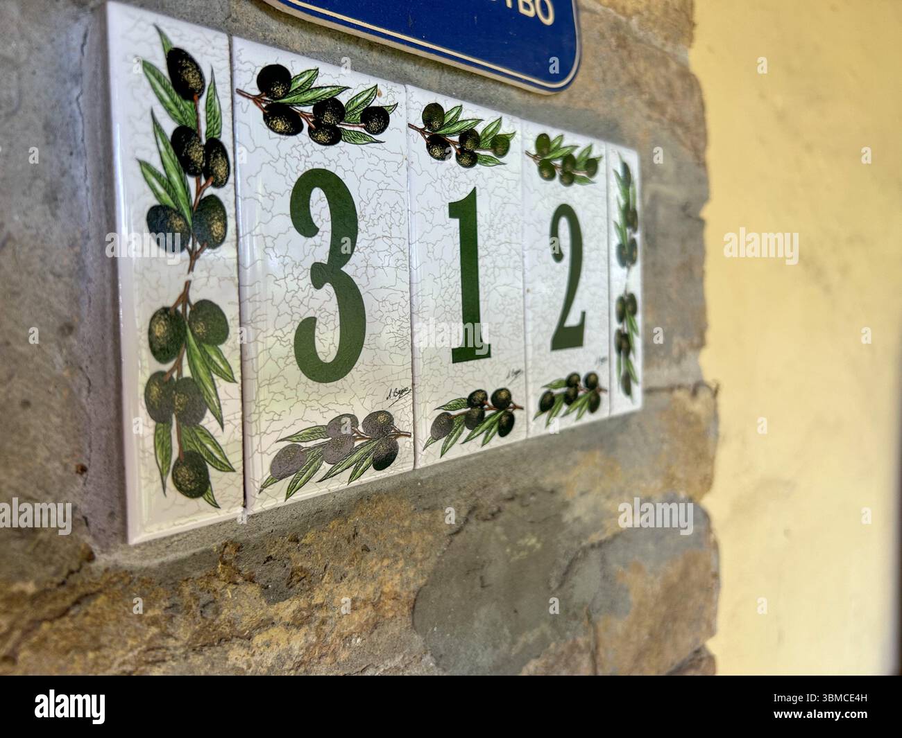 Decorative ceramic house number plate displaying “312” with hand-painted olive branches, mounted on a stone wall next to a rustic yellow facade. - Smartphone Captured Stock Image