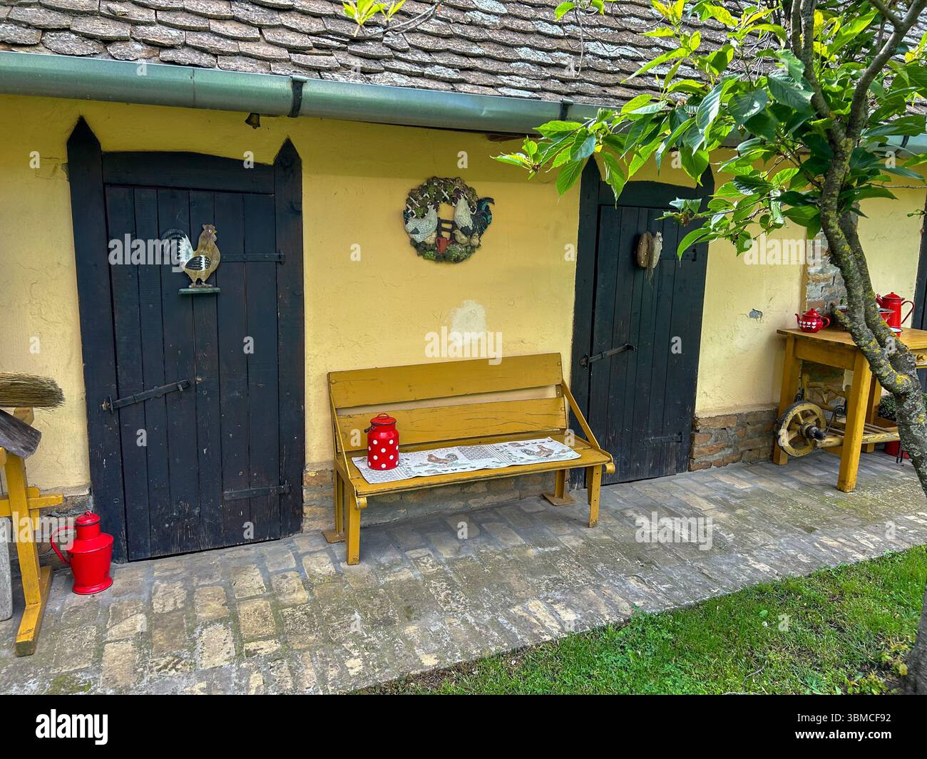 Charming farmhouse exterior with dark wooden doors, rooster decorations, a yellow bench, and red vintage kitchenware creating a cozy countryside vibe. - Smartphone Captured Stock Image