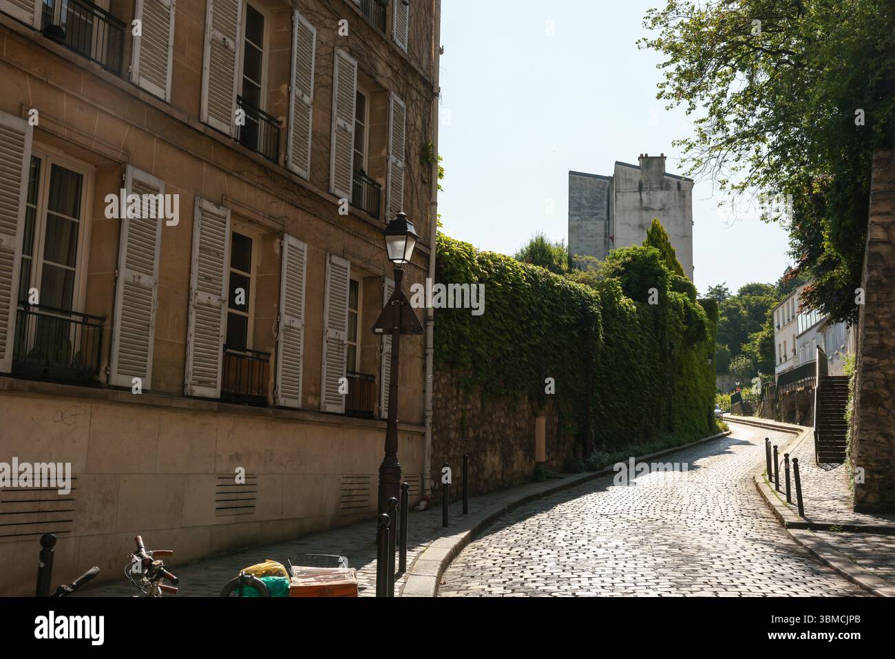 A charming Parisian street scene featuring a cobblestone road, an old beige building with white shutters, and a lush ivy-covered wall. The sunlight ca Stock Photo