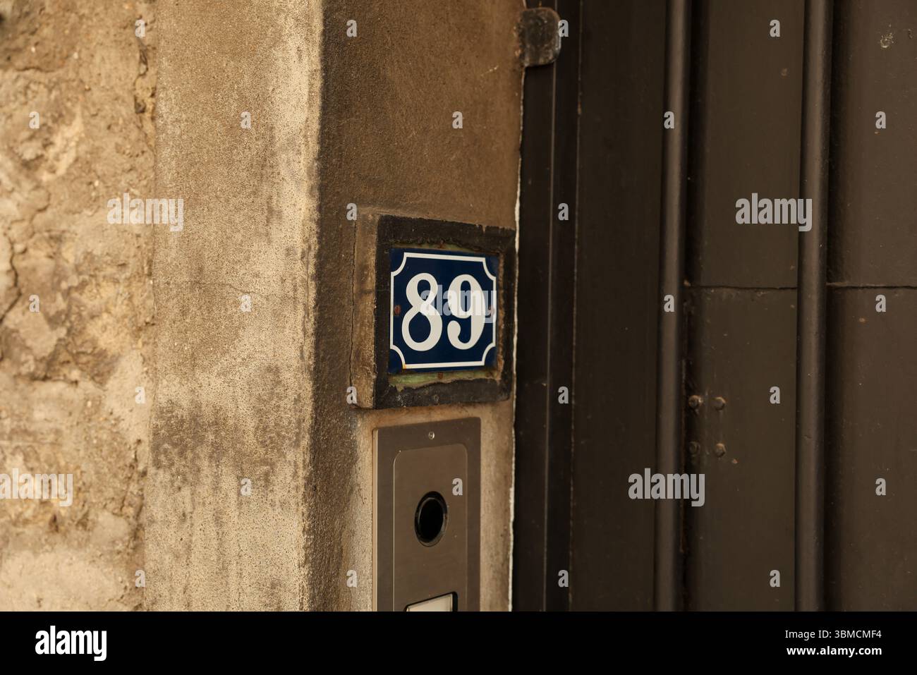 A close-up shot of a weathered building exterior featuring a blue enamel house number eighty-nine affixed to a worn stone wall next to a dark brown Stock Photo