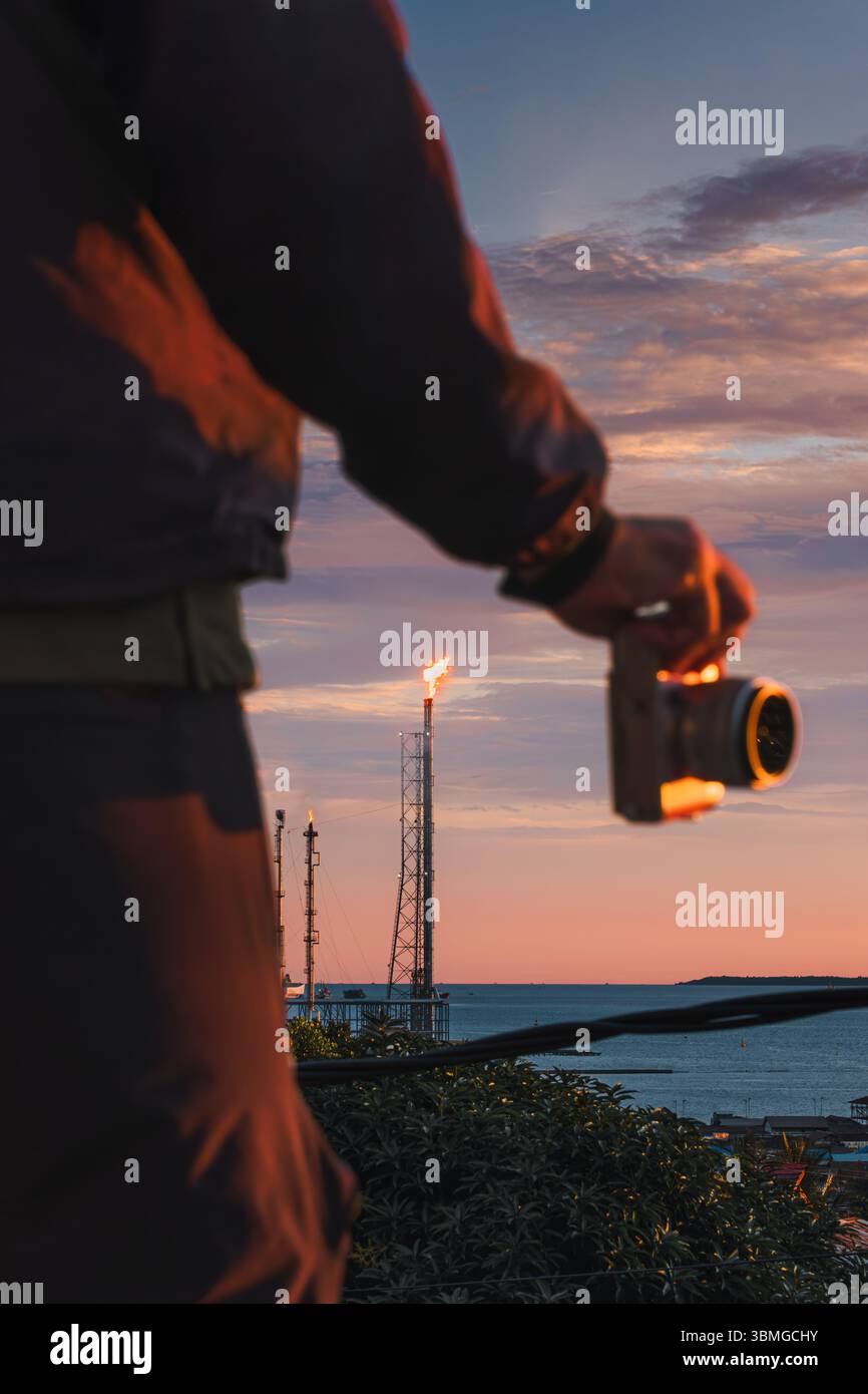 Photographer holding camera in foreground with oil refinery flare burning at sunset, viewed from Gunung Gembira Hill, Balikpapan, East Kalimantan, Ind Stock Photo