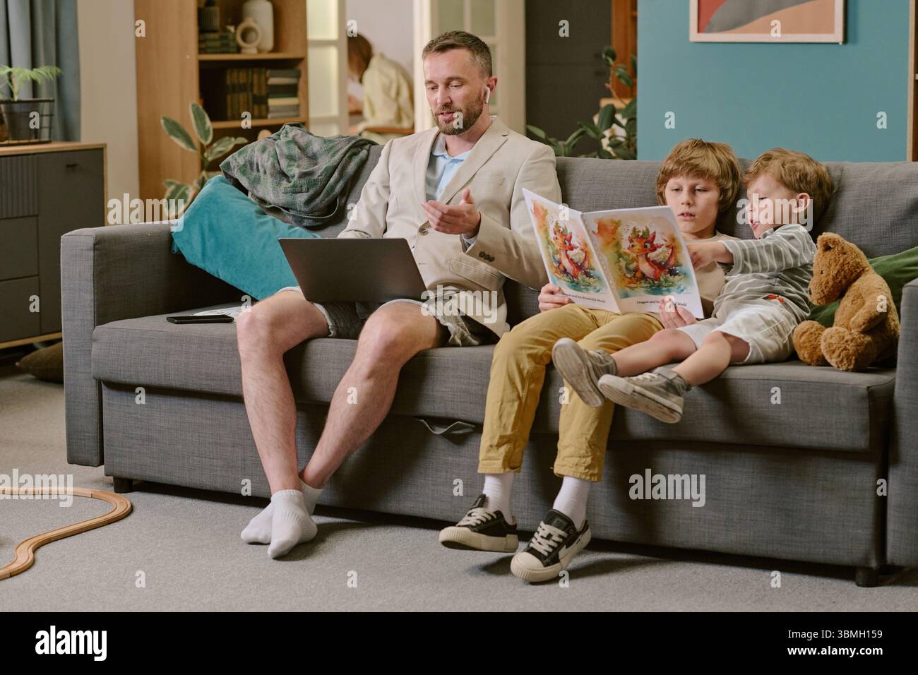 Middle Aged Man Working Remotely While Sons Reading Storybook Stock Photo