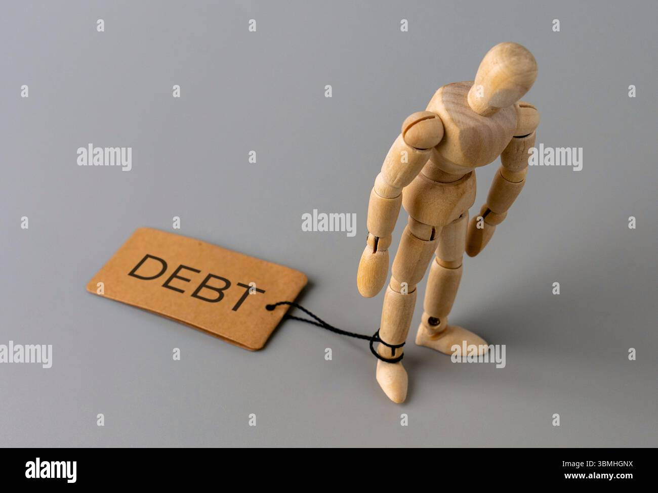 A wooden figure stands with one ankle tied by a string to a tag labeled "DEBT". Financial burden, obligation, being tied down by debt Stock Photo