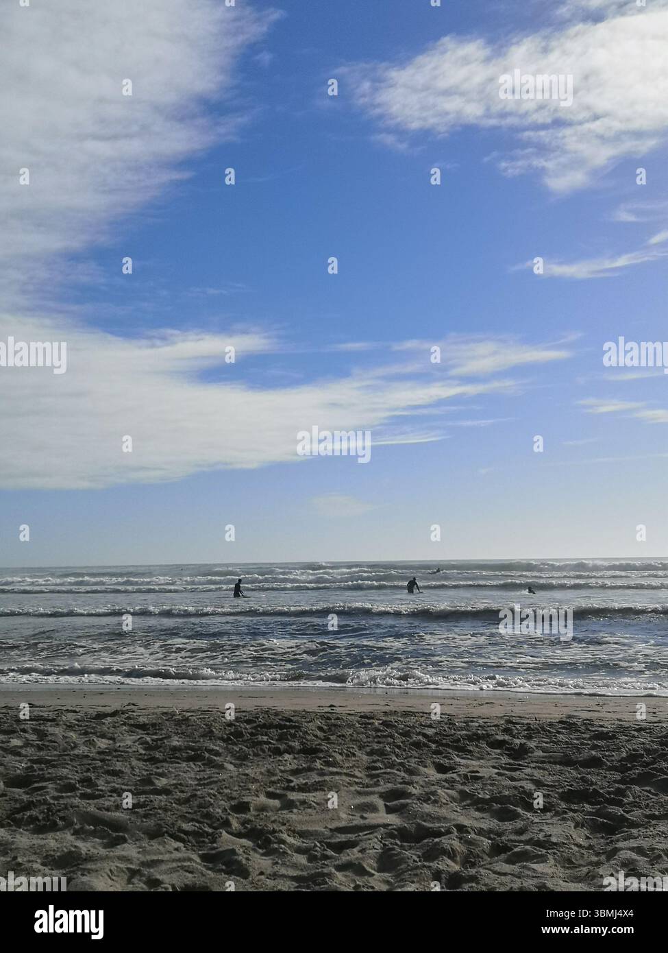 Sandy beach with surfers in the water under a partly cloudy blue sky, capturing a peaceful coastal scene on the Atlantic Ocean. - Smartphone Captured Stock Image