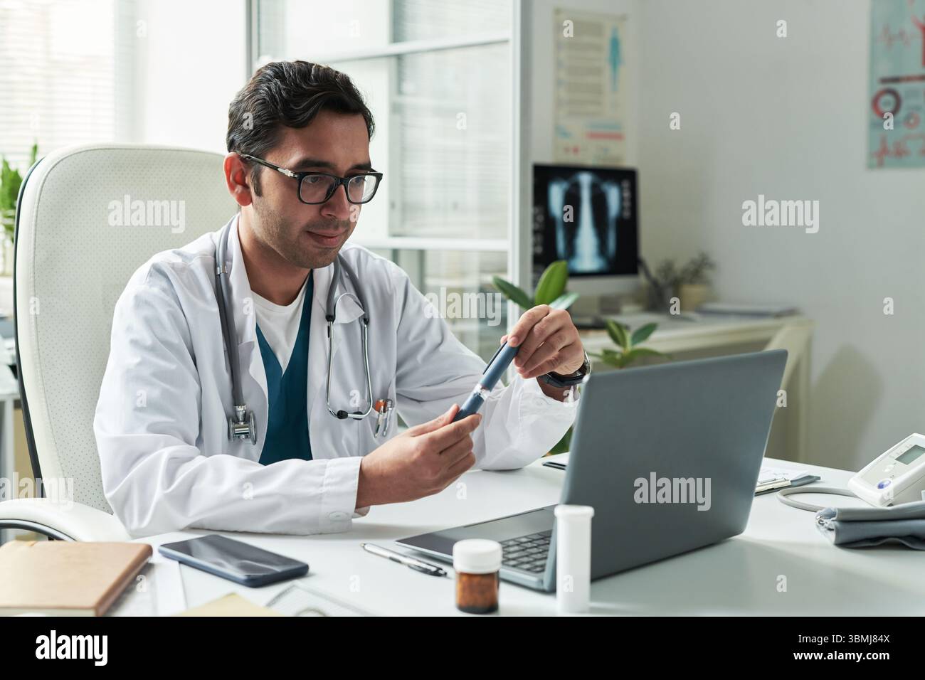 Middle aged Caucasian male doctor sitting at desk examining insulin pen while consulting diabetic patient in medical office, laptop and medication bot Stock Photo