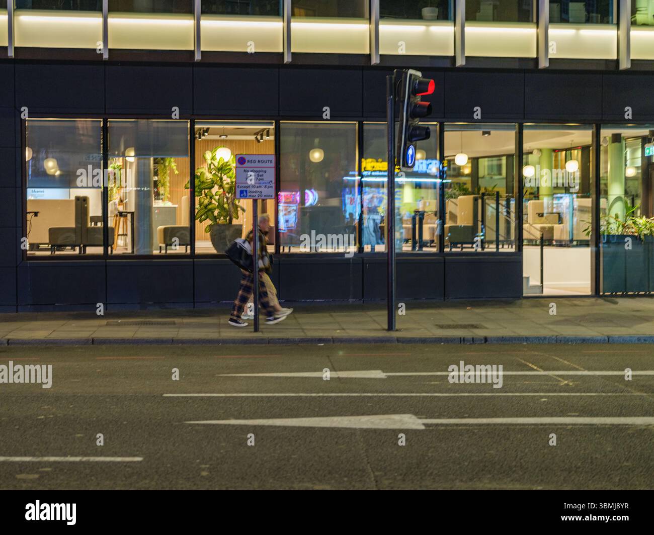 United Kingdom, London, 30 Jun 2025. Couple walking at night past a brightly lit café in central London, with a red pedestrian light and reflections i Stock Photo
